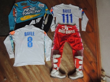 Une vue de dos de maillots de JMB 1989, 1991 et 1992 mais aussi un pantalon Bad bayle de 1989 et une paire de bottes